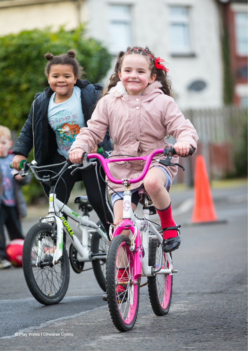 Two young girls cycling happily towards the camera with a traffic cone in the background showing that the street has restricted car access.
