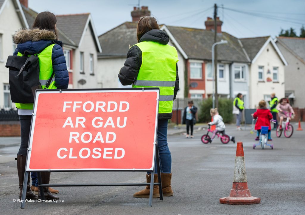 Road closure sign with adults in high viz jackets standing behind it and a traffic cone restricting car access to allow children to play safely on the street where they live