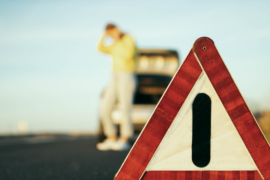 in the foreground is a red-white close-up warning triangle, in the background a girl in a yellow sweater and jeans stands with her hand raised, speaks on the phone, stops the car for roadside assistance, upset. Car breakdown on the road