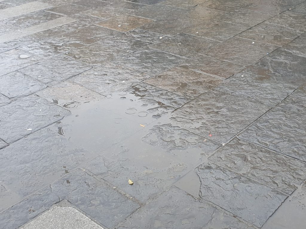 Photo of grey and pale brown coloured paving slabs that have been recently rained on with a puddle collecting on 2 paving slabs in the centre left of the image.