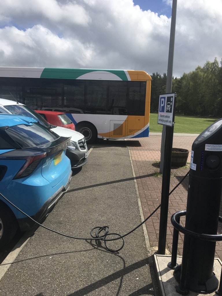 Colour photo of a blue car parked in a car park with other cars and a bus also parked going off into the distance, with an electric car charging cord laying across the pavement between the blue car and the charging point, causing a trip hazard and barrier to pedestrians. 