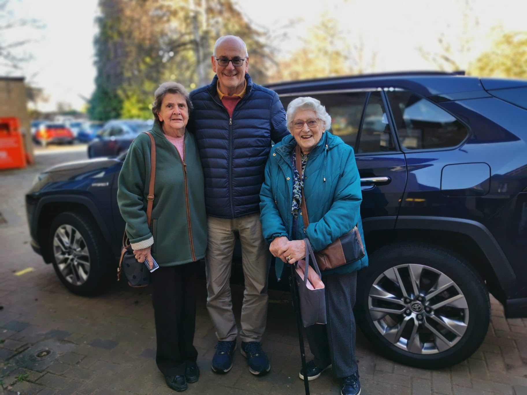 photo of older man with glasses standing between two older ladies, one with a walking stick, all standing next to a blue car.